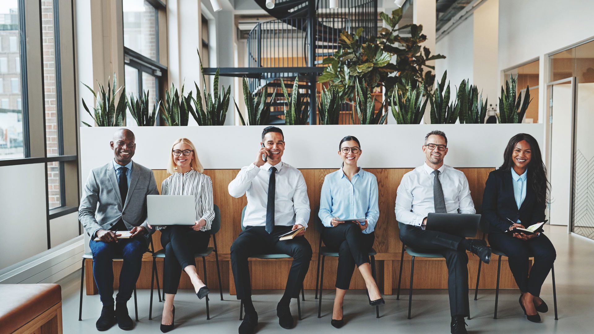 men and women waiting in chairs at an office with computers and notepades